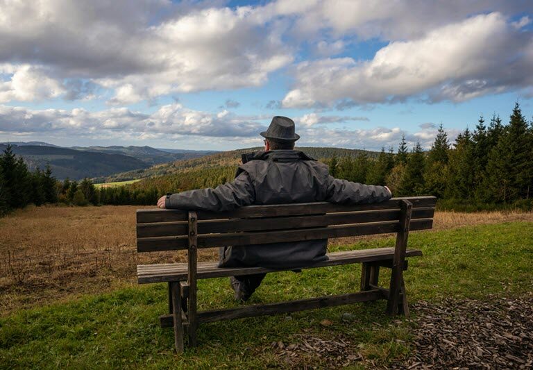 Older man sitting alone on a bench looking out to a beautiful view with dark clouds above
