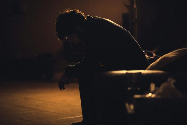 person in a dark room leaning forward in their chair with their head in their hands