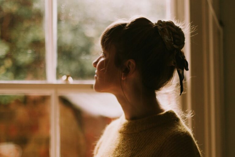 Woman standing alone by a window holding a cup of tea, looking thoughtful