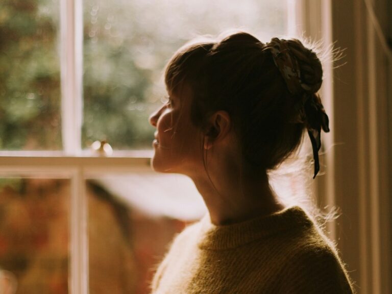 Woman standing alone by a window holding a cup of tea, looking thoughtful